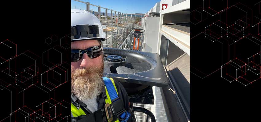 Man in hard hat and sunglasses in front of cooling unit outside.